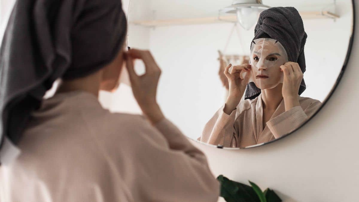 Woman in a robe and hair towel applying a white sheet mask in a round bathroom mirror for a self-care routine.