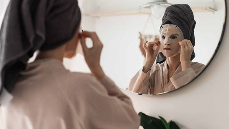 Woman in a robe and hair towel applying a white sheet mask in a round bathroom mirror for a self-care routine.