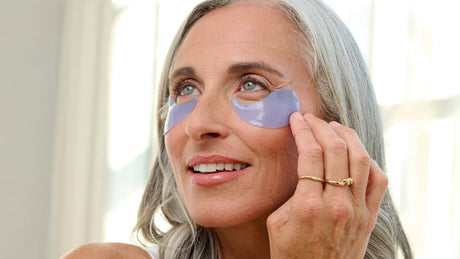 Mature woman applying purple hydrogel under-eye patches, gently pressing them into place to target puffiness and fine lines for a refreshed, hydrated look.