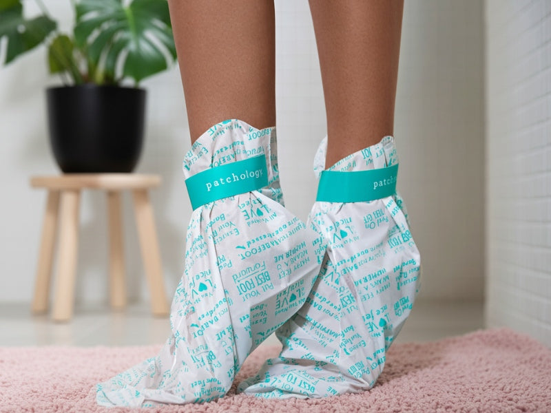 Feet wearing Patchology hydrating foot mask booties on soft rug in bathroom for moisturizing treatment