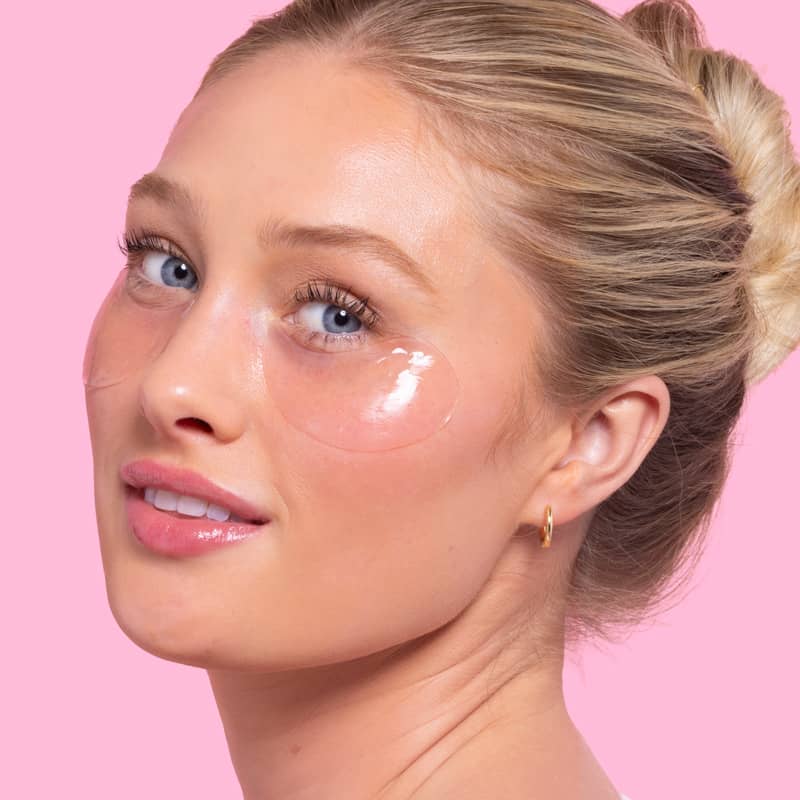 Woman applying rose hydrating eye patches under her eyes with a bright pink background