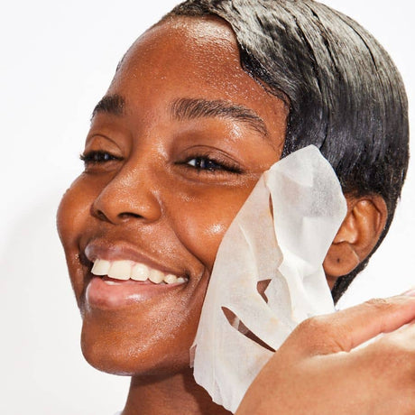 A smiling woman with glowing skin peeling off a Patchology sheet mask to reveal a hydrated and refreshed complexion.